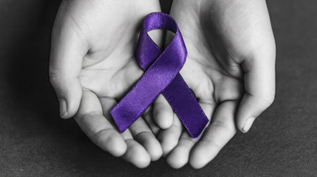 A pair of hands cradles a vibrant purple ribbon against a soft grayscale backdrop. The image displays a close-up composition with selective color focus, highlighting the ribbon's texture and form. Suitable for various editorial content or design projects conveying support, raising awareness, or representing causes.の素材