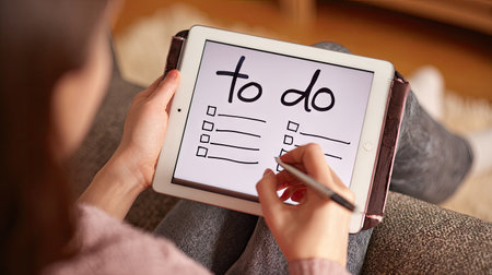 A person is using a tablet to write on a to-do list, emphasizing planning and organization. The scene is likely indoors, featuring a neutral background with the tablet being the central focus. The composition uses shallow depth of field and soft lighting. Suitable for commercial and editorial content.の素材