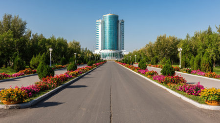 A wide asphalt road leads towards a tall modern building with glass windows under a bright blue sky. The scene features vibrant flowerbeds and lush greenery on either side of the pathway. The image showcases a clear day, possibly suitable for architectural, promotional, or travel related content.の素材
