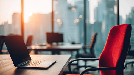 A laptop rests on a desk in a modern office, with a vibrant red chair in the foreground. The image features a large window overlooking a cityscape with sunlight streaming through. The composition suggests a corporate or professional environment suitable for business and commercial purposes.の素材