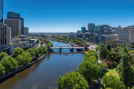 An aerial perspective showcases a river flowing through a city, with a bridge spanning across. The scene features lush green trees lining the riverbanks. Buildings of various heights create an urban backdrop under a clear blue sky. This image may be suitable for architectural, travel, or commercial uses.の素材