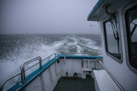 A boat navigates rough ocean waters, leaving a foamy wake behind. The side view shows a portion of the vessel's structure, painted in white and blue. The overcast sky and stormy sea create a somber atmosphere. This image is suitable for various commercial uses, including travel and maritime-related themes.の素材