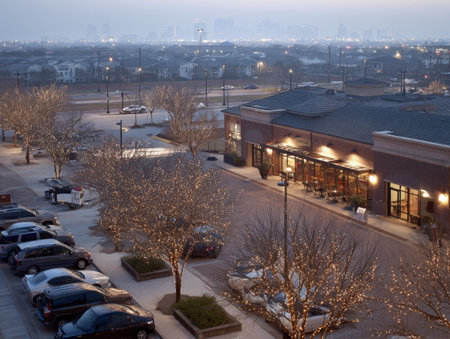 An aerial view presents a commercial area during dusk. Buildings with illuminated storefronts stand beside parking areas occupied by vehicles. Bare trees line the streets, and soft lighting illuminates the scene. Suitable for general business, urban design, or lifestyle publications and projects.の素材