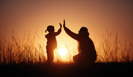 A silhouette depicts a father and child celebrating with a high-five gesture. The image features warm orange and yellow tones from a setting sun, highlighting the figures against the sky. The composition emphasizes the relationship between the two figures, suitable for illustrating family dynamics or related concepts.の素材