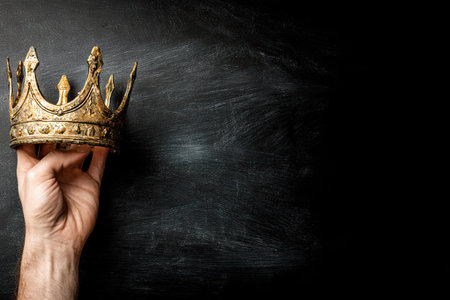 A hand holds a gold crown against a dark, textured backdrop. The composition features strong contrast with warm light highlighting the crown's details. The image evokes themes of royalty and power. Suitable for a variety of projects, it lends itself well to editorial and commercial applications.の素材