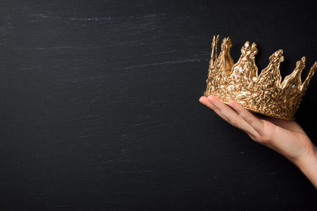 A hand delicately holds a gold-colored crown against a dark, featureless background. The image presents the crown in sharp focus with a textured surface, showcasing its ornate design. The lighting appears soft, accentuating the metallic sheen. This image is suitable for various commercial uses, including representing authority or success.の素材