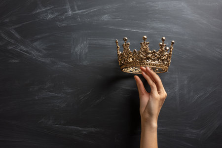 A hand delicately holds a gold crown against a dark, textured backdrop. The composition features a close-up perspective with a focus on detail and contrast. This image displays a luxurious feel with its rich color palette and highlights the concept of power, royalty, and success and could be used in various commercial or editorial projects.の素材