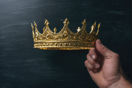 A close-up captures a golden crown held by a hand against a dark, textured background. The crown, highly detailed, shines with a metallic sheen, suggesting wealth and power. The composition highlights the crown, using contrast and focus. This image is suitable for concepts of leadership, success, or royalty in various media.の素材