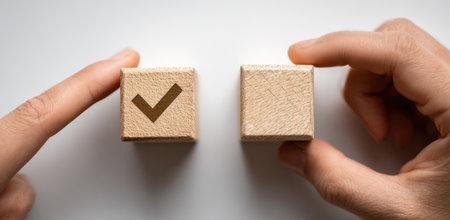 Two hands are featured, holding wooden cubes with a check mark symbol. The cubes are positioned against a plain, white backdrop, casting soft shadows. The image showcases simple elements, suitable for various uses, including marketing and communication purposes. The composition uses a flat lay design for visual simplicity.の素材