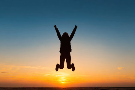 A silhouette of a person jumping with arms raised is set against a vibrant gradient sky. The scene features warm oranges and yellows blending into blue. The composition emphasizes freedom and accomplishment. This image is suitable for various commercial uses, including advertising and promotional materials.の素材