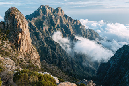 Dramatic view of mountain peaks against a cloudy sky. The image showcases textures of rock and vegetation under natural light. The composition highlights the high altitude with clouds filling the valleys, suggesting vastness and inspiring awe. Suitable for editorial and commercial use.の素材