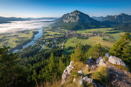 An aerial view presents a mountain landscape featuring a river snaking through a valley. A town is nestled amidst green fields and forests. The scene is illuminated by natural sunlight under a clear, blue sky. This image is suitable for various commercial uses, illustrating natural beauty and outdoor environments.の素材
