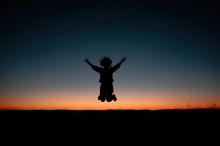 A silhouette of a person jumping in the air, arms raised, against a vibrant sunset sky. The composition emphasizes the contrast between dark and bright colors. The image suggests concepts of freedom and joy, suitable for various editorial and commercial applications. The lighting is low-key, with a strong backlighting creating the silhouette.の素材