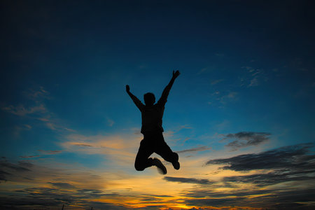 A silhouette depicts a person mid-jump with arms raised against a vivid sunset. The sky transitions through shades of blue and orange, suggesting an outdoor environment. This image utilizes a silhouette technique, creating a dramatic effect. Suitable for various projects needing visuals related to freedom or achievement.の素材