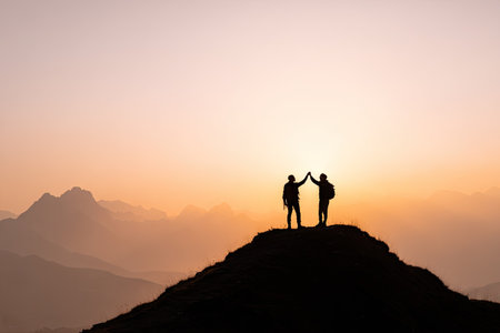 Two figures stand atop a mountain, silhouetted against a gradient sky of oranges and pinks. They appear to be celebrating, arms raised in a gesture of achievement. The lighting suggests either sunrise or sunset, creating a warm, inviting atmosphere. This image could be used for various purposes, including motivational or inspirational content.の素材