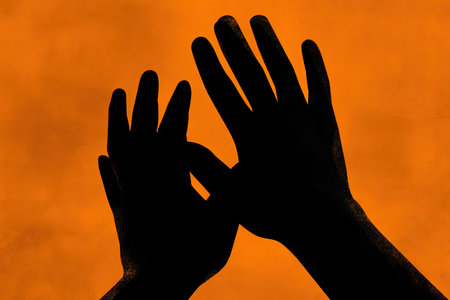 Two hands are silhouetted against a bright orange backdrop. The composition shows the hands in a reaching or gesturing position. The lighting creates strong contrast highlighting the hand's shape. This image could be suitable for various commercial purposes, including illustrating concepts or design elements.の素材