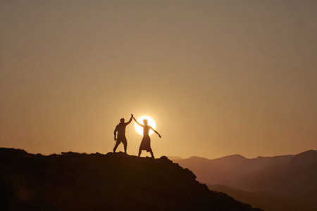 Two individuals stand atop a mountain ridge, their silhouettes dramatically framed by a setting sun. Warm tones dominate the scene, with the sky displaying gradients of orange and yellow. The image features a clear, overhead composition, ideal for illustrating concepts such as achievement or collaboration. Suitable for various commercial and editorial applications.の素材