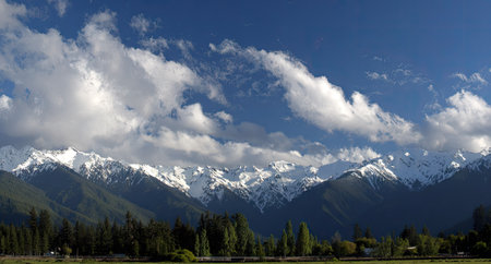An image captures a panoramic view of majestic mountains with snow-covered peaks set against a clear, blue sky. Fluffy white clouds drift above, creating a visually appealing contrast. The foreground is filled with lush greenery. This scene is suitable for various commercial uses.の素材