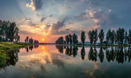 A scenic landscape showcases a lake reflecting the sky at sunset. The composition includes trees along the shoreline and a dramatic sky with clouds and sun rays. The image's lighting creates a sense of serenity. Suitable for diverse commercial and editorial purposes.の素材