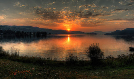 A stunning sunset illuminates a serene lake, reflecting warm orange and yellow hues. Mountains form a dark silhouette against the vibrant sky. The scene is bathed in soft, natural light, creating a peaceful ambiance. Suitable for a variety of editorial and commercial applications.の素材