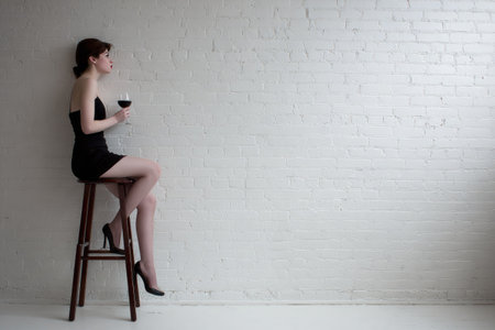 A woman in a short black dress is seated on a tall stool, holding a wine glass. The minimalist composition features a white brick wall as a backdrop. The lighting is soft, highlighting the woman's form. This image is suitable for various commercial purposes, including editorial content.の素材