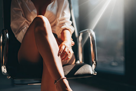 A woman sits in a chair, with sunlight streaming across her figure. The image emphasizes the legs and torso of the subject, with a focus on light and shadow. The scene takes place indoors, potentially in a modern office or domestic space. Suitable for various commercial or editorial applications.の素材