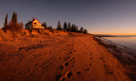 Warm, golden-hour shot of a small house on a sandy beach, footprints, trees, &amp; calm waterの素材