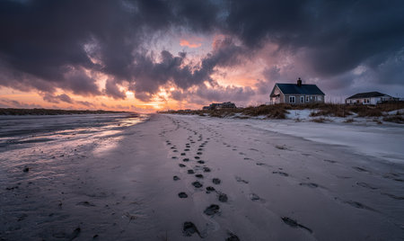 Footprints lead across a beach towards a house as a vibrant sunset paints the skyの素材