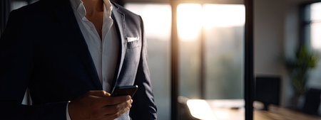 A man in a formal suit stands near a window, illuminated by sunlight. The image showcases a professional attire in a modern office setting. The composition focuses on the upper body, emphasizing a confident posture. Suitable for various business, communication, and professional concept use.の素材