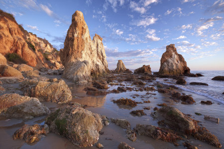 Dramatic coastal scene showcasing large rock formations near water at sunset. Warm tones dominate the landscape, with golden rock surfaces contrasting against the blue sky and water. The composition highlights reflections and shadows, suggesting a serene yet powerful environment suitable for various commercial purposes.の素材