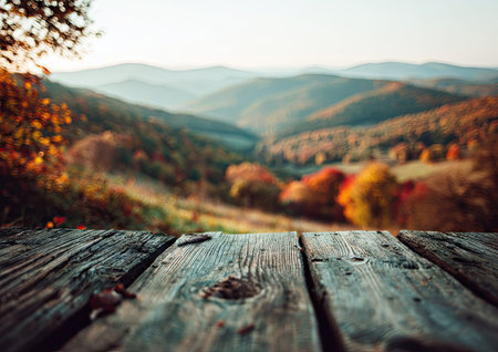 A close-up of a weathered wooden table leads to a blurred view of a fall landscape. The color palette features warm tones of orange and brown, complemented by soft green hues of trees and hills. The natural light suggests a day environment, suitable for various promotional and advertising applications.の素材