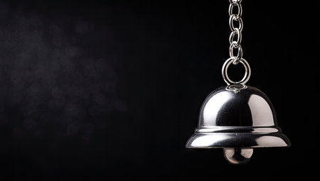 A close-up showcases a metallic bell suspended from a chain, set against a dark backdrop. The bell reflects light, displaying a polished surface. The composition creates a sense of depth, with subtle shadows and diffused lighting. Suitable for advertising and illustrative projects, this image offers copy space for various creative applications.の素材