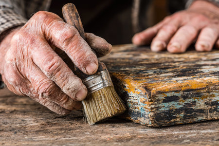 An aged craftsman's hands hold a brush over a weathered wooden surface, possibly preparing it for a new coat. The composition highlights textured skin and a well-used brush. The image features warm tones and a shallow depth of field, suggesting interior lighting, potentially suitable for editorial content or commercial projects.の素材