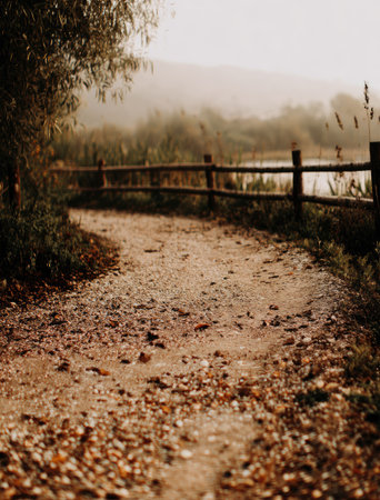 A winding dirt path leads towards a wooden fence, hinting at a body of water or landscape. The image displays a natural scene with warm colors, featuring tones of brown, and green. The composition highlights a sense of tranquility, which could be used for various editorial or commercial projects.の素材