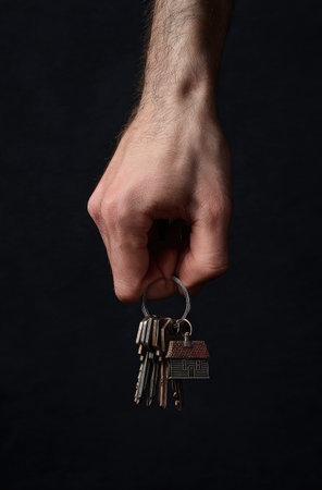 A hand holds a set of keys, presented against a stark, black backdrop. The image displays a close-up perspective, highlighting textures and shapes. The lighting is focused, creating shadows and enhancing the contrast. This could be used for various projects related to real estate, security, or access.の素材