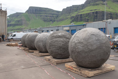 Six large stone spheres rest on wooden pallets outdoors near industrial structures and a mountainous landscape. The gray stones exhibit a textured surface, contrasting with the smooth textures of the buildings and surrounding environment. This scene, under overcast lighting, might be suitable for illustrating construction or geological themes for various commercial applications.の素材