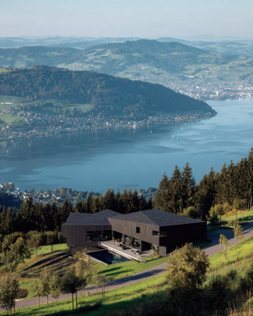 A contemporary architectural design features a dark-colored residence nestled on a hillside. The structure showcases clean lines and geometric forms. The scene captures a daytime view, with the house overlooking a vast lake and distant mountains. This image is suitable for architectural, travel, or lifestyle content, and various editorial needs.の素材