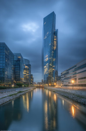 A tall, modern skyscraper dominates the image, mirrored in the calm water below. The building's glass facade reflects the cloudy sky and surrounding buildings. The composition features a symmetrical view with soft lighting and a muted color palette, possibly suitable for architectural or urban design projects.の素材
