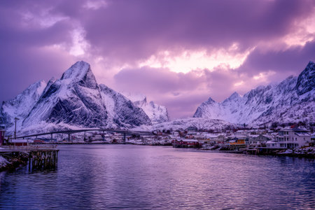 This landscape image features snow-covered mountains reflected in calm water. The composition is highlighted by a vibrant purple sky, with fluffy clouds. The scene is illuminated by soft light. It may be suitable for a range of purposes including editorial content and website design.の素材