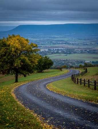 A scenic winding road weaves through a landscape dominated by green fields, trees with yellow leaves, and a distant mountain range. The overcast sky suggests a day with diffused lighting, and a fence lines one side of the road. This image could be used for travel, nature, or environmental themes.の素材