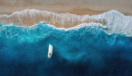 An overhead view reveals a small boat in vibrant blue water near a beach with foamy waves. The image displays a contrasting mix of sandy beige, white foam, and deep blue hues. Suitable for commercial applications, the image offers visual storytelling possibilities related to travel and the natural world.の素材