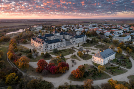 An aerial photograph showcases a large building with multiple wings and a patterned roof. Surrounding the structure are trees, some displaying vibrant autumn colors. The composition is set against a town landscape under a sunset sky, with soft lighting enhancing the scene. This image may be suitable for architectural, travel, or historical themes.の素材