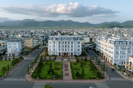 An aerial perspective captures multiple large buildings with light-colored facades. Roads and green spaces surround the structures, while the sky features clouds. The image displays a balanced composition. It could be useful for real estate, urban planning, or architectural projects.の素材