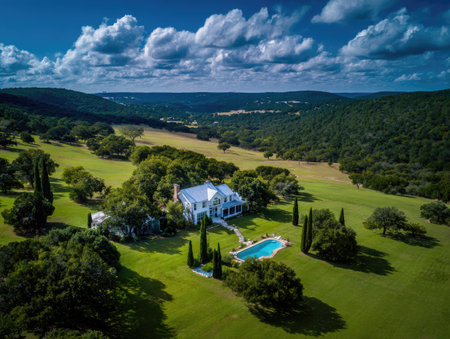 An aerial perspective showcases a large house with a swimming pool situated within a vast, green landscape. The image displays a bright, sunny day with a vibrant blue sky dotted with clouds. The composition offers an expansive view of rolling hills and dense foliage, suitable for various editorial and commercial applications.の素材