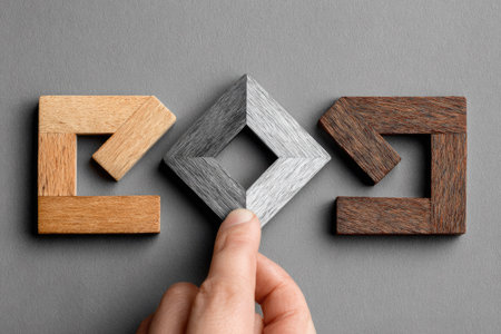 An image showcases three geometric wooden puzzle pieces in shades of brown and gray on a gray background. A hand is seen interacting with the central piece. The composition features overhead lighting and a close-up perspective, suggesting a focus on texture and form. Suitable for illustrating concepts of design and problem-solving.の素材