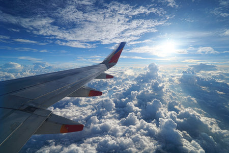 An airplane wing extends into a vibrant sky filled with puffy white clouds. The composition features the wing's metallic surface, contrasted against the bright sun and deep blue hues. This aerial view suggests a sense of travel and transportation, suitable for various commercial uses.の素材