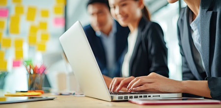 An image shows three people engaged in work using a laptop computer. They are dressed in business attire, working in a modern office environment. The composition features soft focus and natural lighting. This image could be used for articles, websites, and marketing materials related to business.の素材