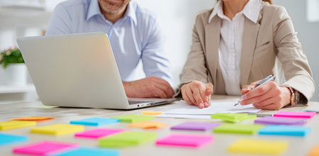 Two individuals are seen collaborating in a well-lit indoor environment. One person uses a laptop, while the other writes on paper. The scene displays a variety of colorful sticky notes. The image suggests teamwork and is suitable for illustrating business, project management, or educational content.の素材