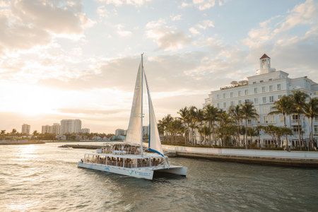 A catamaran sails across the water on a sunny day. The vessel is white with white sails. A large building and a distant skyline are visible in the background, set against a partly cloudy sky. This image is suitable for various commercial uses, including travel and lifestyle content.の素材