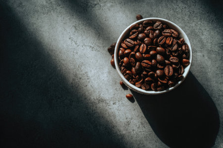 A close-up captures a white bowl filled with roasted coffee beans. The image showcases rich brown tones and textural details of the beans. Dramatic lighting casts shadows across a textured surface. The composition suggests potential use for culinary, beverage, or lifestyle-related projects.の素材